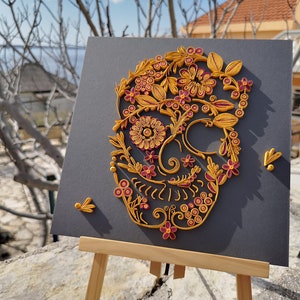 K&ouml;nnte beinhalten: Ein Papier-Quilling-Kunstwerk, das einen Sch&auml;del mit aufwendigen floralen Mustern in Gelb- und Rosat&ouml;nen auf einem grauen Hintergrund zeigt.