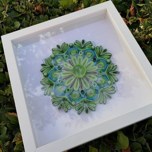 May include: A framed green and blue paper quilling mandala art piece. The mandala is a circular design with intricate details and is framed in a white frame.