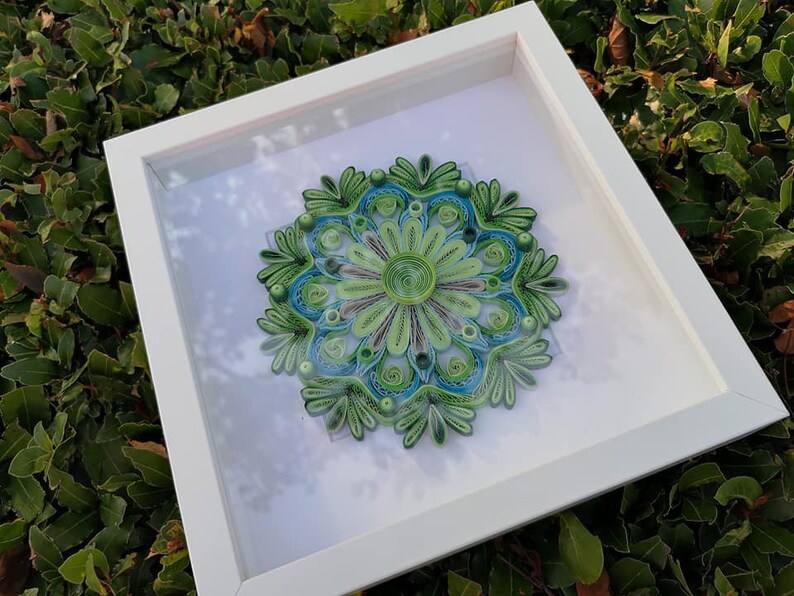 May include: A framed green and blue paper quilling mandala art piece. The mandala is a circular design with intricate details and is framed in a white frame.