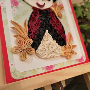 May include: A handmade paper quilling art piece featuring a doll with a red, black, and white dress. The doll is surrounded by flowers and leaves made of paper.