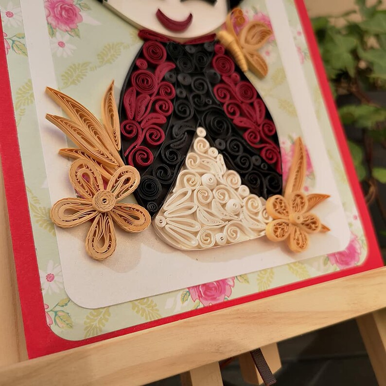 May include: A handmade paper quilling art piece featuring a doll with a red, black, and white dress. The doll is surrounded by flowers and leaves made of paper.