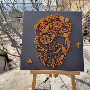 K&ouml;nnte beinhalten: Ein Papier-Quilling-Kunstwerk eines Sch&auml;dels mit floralen Details in Gold und Burgunderrot auf grauem Hintergrund. Das Kunstwerk wird auf einem Holzstaffelei ausgestellt.