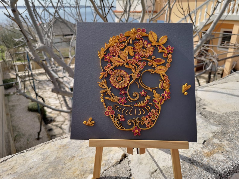 K&ouml;nnte beinhalten: Ein Papier-Quilling-Kunstwerk eines Sch&auml;dels mit floralen Details in Gold und Burgunderrot auf grauem Hintergrund. Das Kunstwerk wird auf einem Holzstaffelei ausgestellt.