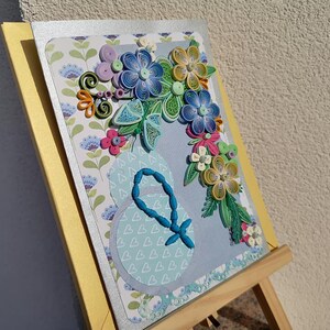 May include: A handmade greeting card with a blue, green, and yellow paper flower bouquet on a light blue background. The card is on a wooden easel.