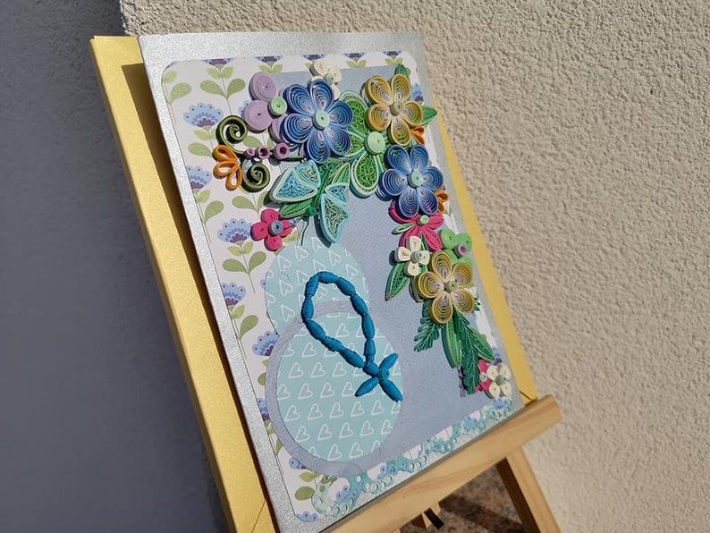 May include: A handmade greeting card with a blue, green, and yellow paper flower bouquet on a light blue background. The card is on a wooden easel.