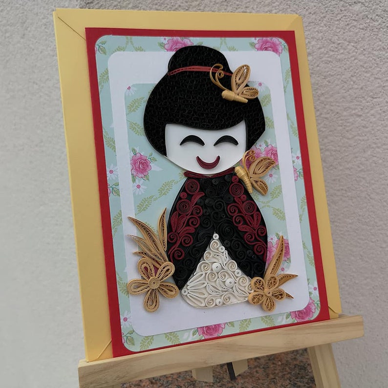 May include: A handmade paper quilling card featuring a smiling geisha doll with black hair, red and black clothing, and white and gold accents. The doll is surrounded by flowers and butterflies.