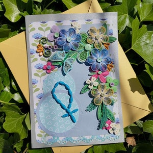 May include: A handmade greeting card with a blue, green, and yellow paper flower design. The card has a blue background and a white border. The card is decorated with a blue rosary.