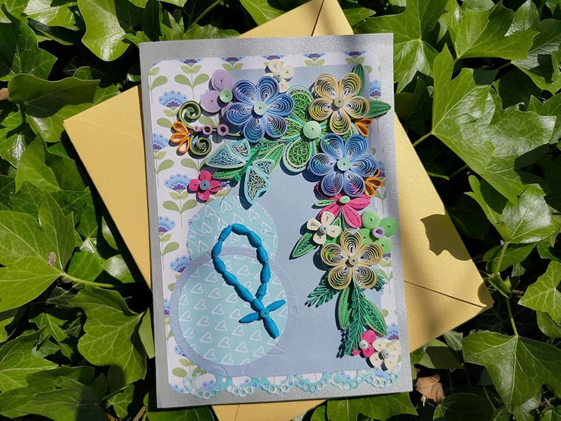 May include: A handmade greeting card with a blue, green, and yellow paper flower design. The card has a blue background and a white border. The card is decorated with a blue rosary.