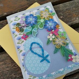 May include: A handmade greeting card with a blue, yellow, and green paper flower design. The card has a blue background with a white heart pattern and a blue butterfly.