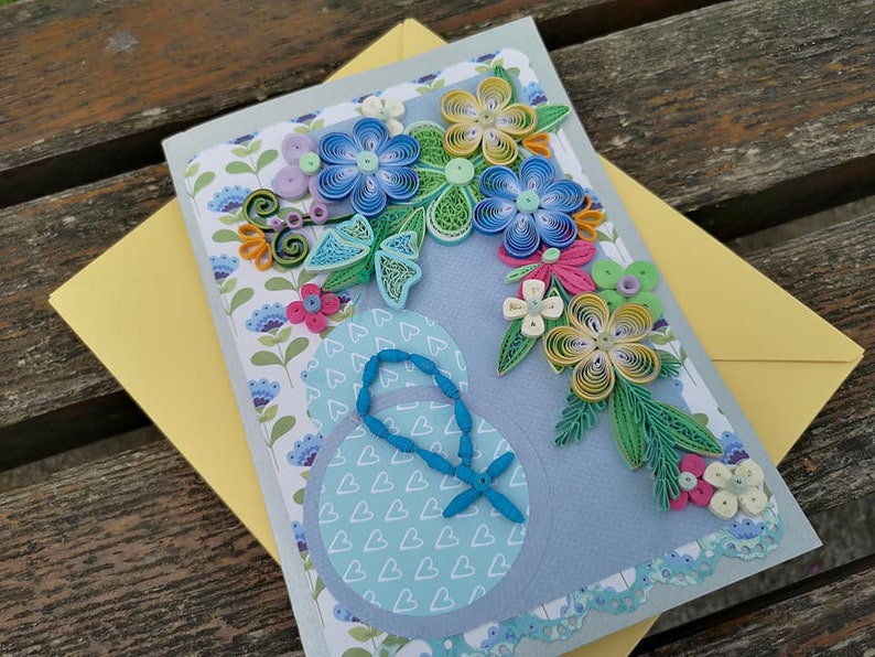 May include: A handmade greeting card with a blue, yellow, and green paper flower design. The card has a blue background with a white heart pattern and a blue butterfly.
