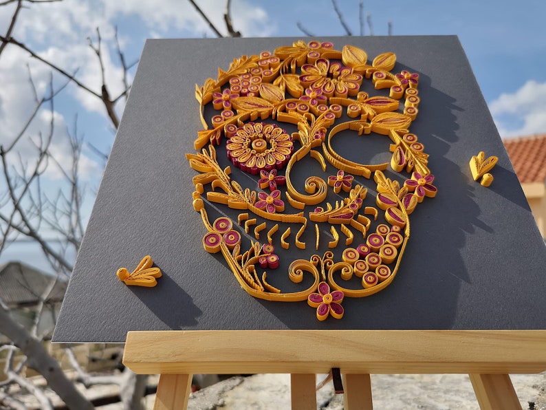 K&ouml;nnte beinhalten: Ein Papierquilling-Kunstwerk eines Zucker-Sch&auml;dels mit gelben und rosa Blumen und Spiralen auf einem grauen Hintergrund.