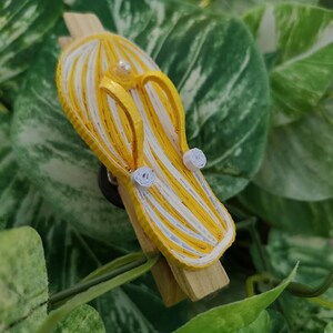 Op de afbeelding: Een gele en witte gestreepte papieren quilling flip-flop met witte papieren quilling bloemen. De flip-flop is bevestigd aan een houten wasknijper.