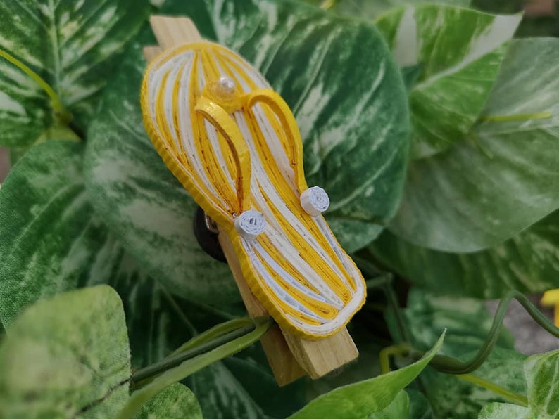 Op de afbeelding: Een gele en witte gestreepte papieren quilling flip-flop met witte papieren quilling bloemen. De flip-flop is bevestigd aan een houten wasknijper.