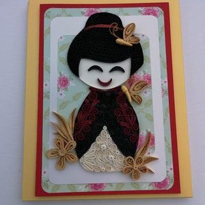 May include: A handmade greeting card featuring a paper quilling art piece of a smiling Japanese doll with a red and black kimono, surrounded by gold and white paper flowers and butterflies on a green and white floral background.