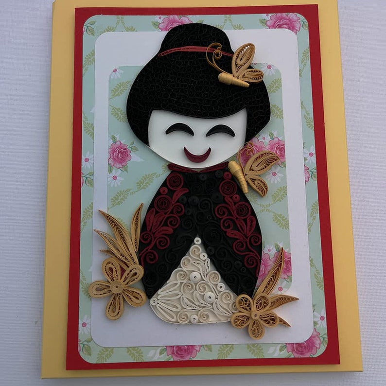 May include: A handmade greeting card featuring a paper quilling art piece of a smiling Japanese doll with a red and black kimono, surrounded by gold and white paper flowers and butterflies on a green and white floral background.