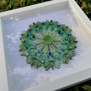 May include: A framed green and blue paper quilling mandala art piece. The mandala is a circular design with intricate details and is framed in a white frame.