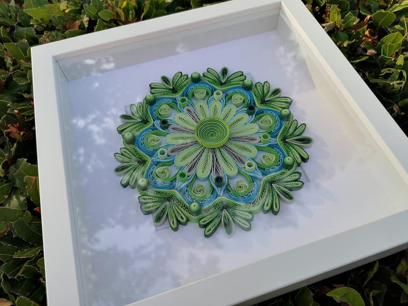 May include: A framed green and blue paper quilling mandala art piece. The mandala is a circular design with intricate details and is framed in a white frame.