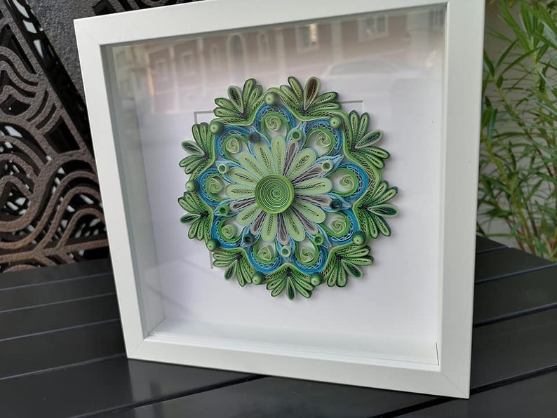 May include: A framed green and blue paper quilling mandala art piece. The mandala is made of paper that has been rolled and shaped into intricate designs.