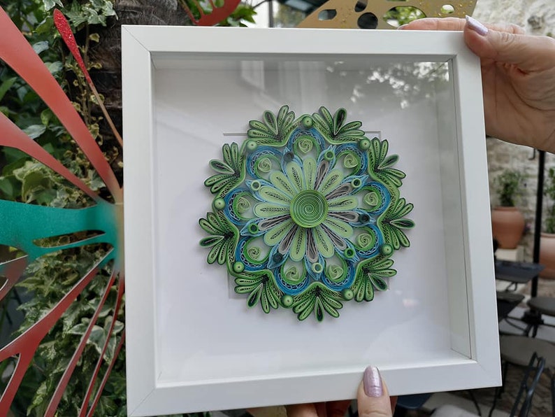 May include: A framed green and blue paper quilling mandala art piece. The mandala is a circular design with intricate patterns and swirls. The frame is white with a glass front.