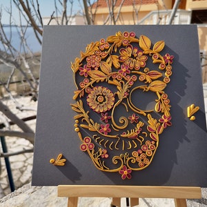 K&ouml;nnte beinhalten: Ein Papier-Quilling-Kunstwerk, das einen Sch&auml;del mit aufwendigen floralen Mustern in Gelb- und Rosat&ouml;nen auf einem grauen Hintergrund zeigt.