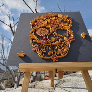 K&ouml;nnte beinhalten: Ein Papierquilling-Kunstwerk eines Zucker-Sch&auml;dels mit filigranen floralen Designs in Gelb- und Burgunder-T&ouml;nen. Das Kunstwerk ist auf einem schwarzen Hintergrund montiert und auf einem Holzstaffelei ausgestellt.