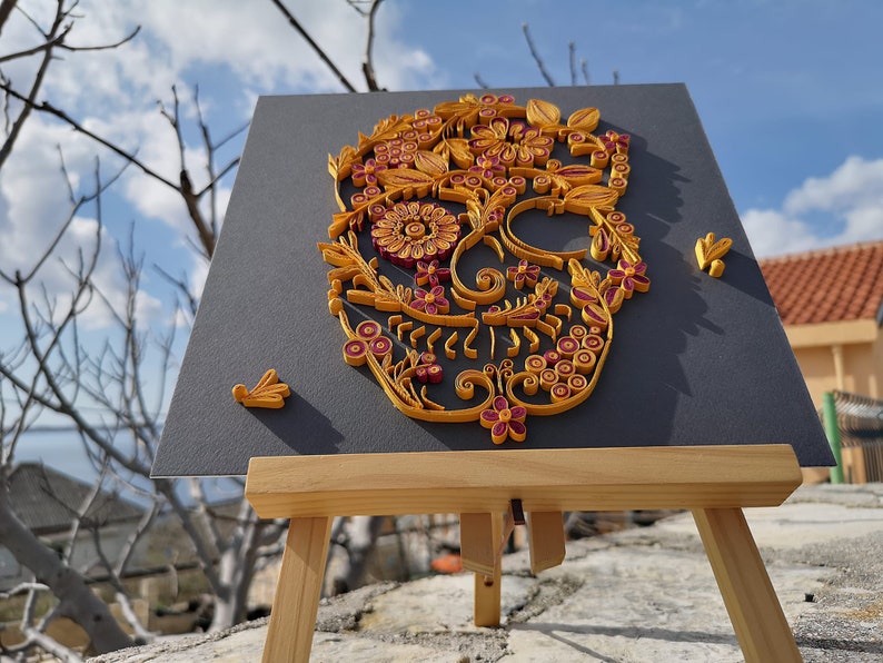 K&ouml;nnte beinhalten: Ein Papierquilling-Kunstwerk eines Zucker-Sch&auml;dels mit filigranen floralen Designs in Gelb- und Burgunder-T&ouml;nen. Das Kunstwerk ist auf einem schwarzen Hintergrund montiert und auf einem Holzstaffelei ausgestellt.