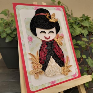 May include: A handmade paper quilling art piece depicting a Japanese doll with black hair, red and black clothing, and white and gold accents. The doll is surrounded by flowers and butterflies.