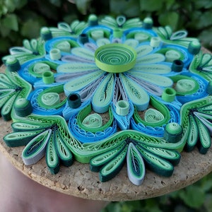 May include: A green, blue, and white paper quilling mandala with a cork backing. The mandala is made of rolled paper strips that are arranged in a circular pattern.