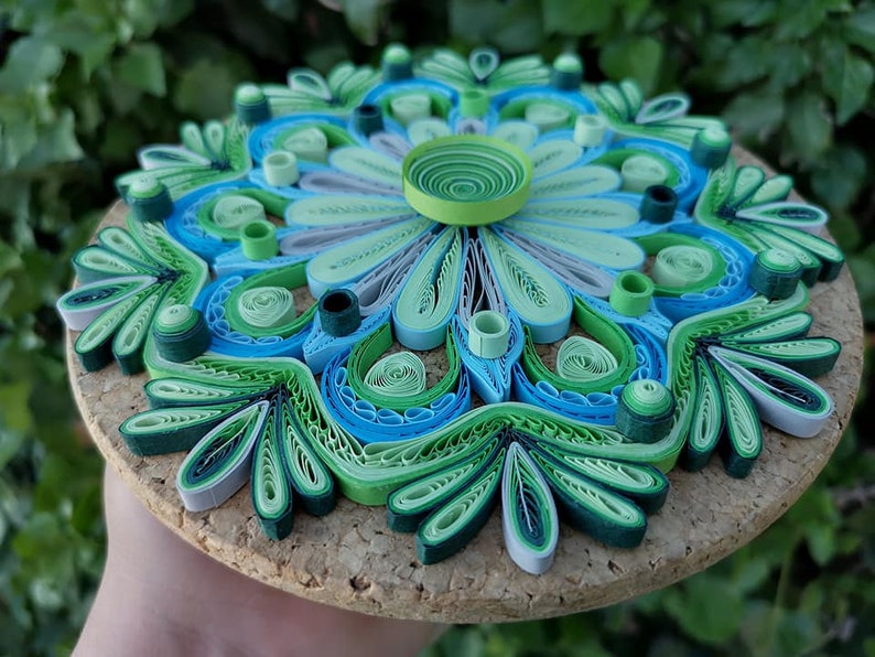 May include: A green, blue, and white paper quilling mandala with a cork backing. The mandala is made of rolled paper strips that are arranged in a circular pattern.