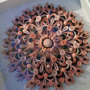 Könnte beinhalten: Ein gerahmtes Papier-Quilling-Kunstwerk mit einem kreisförmigen Design. Das Design zeigt ein Mandala mit filigranen Mustern in Rosatönen, Pfirsich und Schwarz.