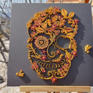 K&ouml;nnte beinhalten: Ein gelbes und pinkfarbenes Papierquilling-Kunstwerk, das einen Sch&auml;del mit floralen Designs darstellt. Der Sch&auml;del besteht aus komplizierten Papierrollen und ist vor einem grauen Hintergrund platziert.