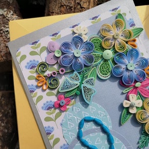 May include: A handmade greeting card with a blue, green, yellow, and pink paper flower arrangement. The flowers are made with quilling, a paper craft technique.