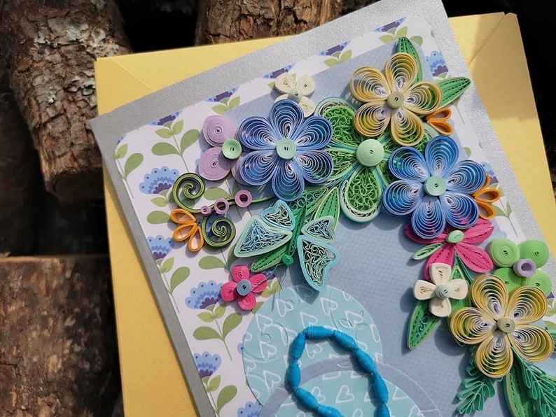 May include: A handmade greeting card with a blue, green, yellow, and pink paper flower arrangement. The flowers are made with quilling, a paper craft technique.