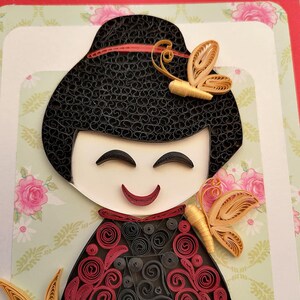 May include: A paper quilling art piece of a smiling woman with black hair and a red and black patterned dress. She is wearing a gold butterfly on her hair. The background is a green and white floral pattern.