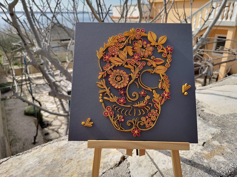 K&ouml;nnte beinhalten: Ein Papierquilling-Kunstwerk aus Gold und Burgunderrot, das einen Sch&auml;del mit floralen Designs auf einem grauen Hintergrund zeigt.