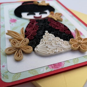 May include: A handmade paper quilling card featuring a smiling doll with black hair, a red and black dress, and white and yellow flowers.