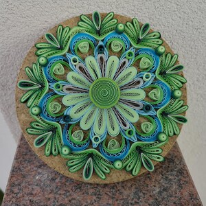 May include: A green, blue, and white paper quilling mandala with a cork backing. The mandala is made of intricate paper rolls and is a beautiful example of paper art.