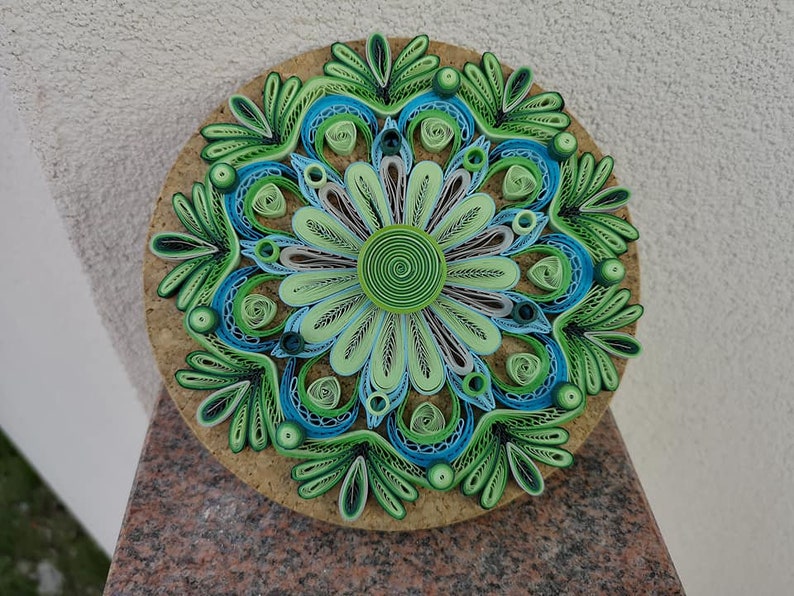 May include: A green, blue, and white paper quilling mandala with a cork backing. The mandala is made of intricate paper rolls and is a beautiful example of paper art.