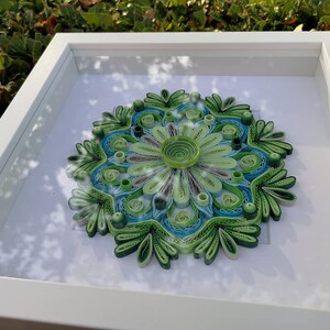 May include: A framed green and blue paper quilling mandala art piece. The mandala is made of paper that has been rolled and shaped into intricate designs.