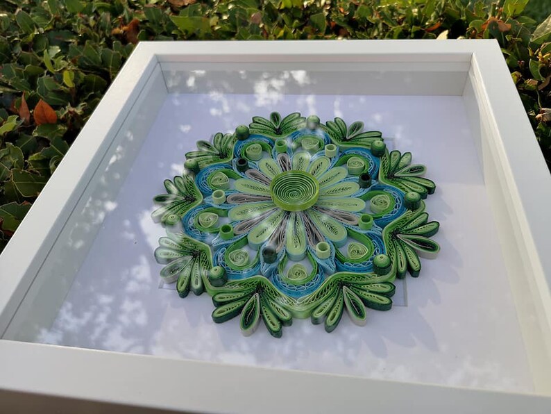 May include: A framed green and blue paper quilling mandala art piece. The mandala is made of paper that has been rolled and shaped into intricate designs.