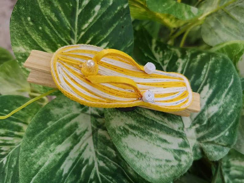 Op de afbeelding: Een gele en witte gestreepte papieren flip-flop met witte parel accenten, bevestigd aan een houten wasknijper.
