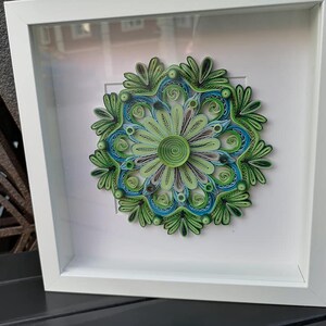 May include: A framed green and blue paper quilling mandala art piece. The mandala is a circular design with intricate details and is framed in a white frame.