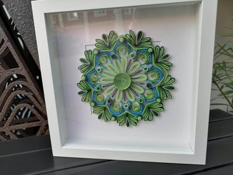May include: A framed green and blue paper quilling mandala art piece. The mandala is a circular design with intricate details and is framed in a white frame.