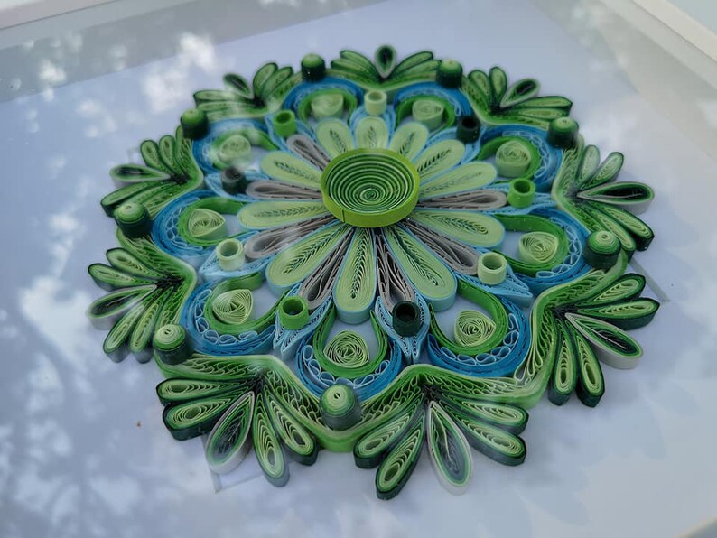 May include: A green, blue, and gray paper quilling mandala with a floral design. The mandala is framed and has a white background.