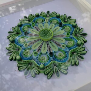 May include: A framed green and blue paper quilling mandala with a white background. The mandala is made of paper that has been rolled and glued together to create a intricate design.