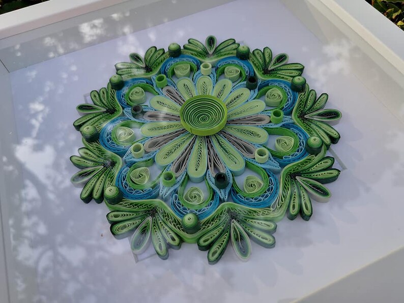 May include: A framed green and blue paper quilling mandala with a white background. The mandala is made of paper that has been rolled and glued together to create a intricate design.