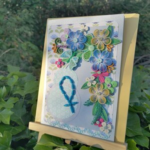 May include: A handmade card with a blue and green paper flower arrangement on a light blue background. The card features a blue rosary and a cross.