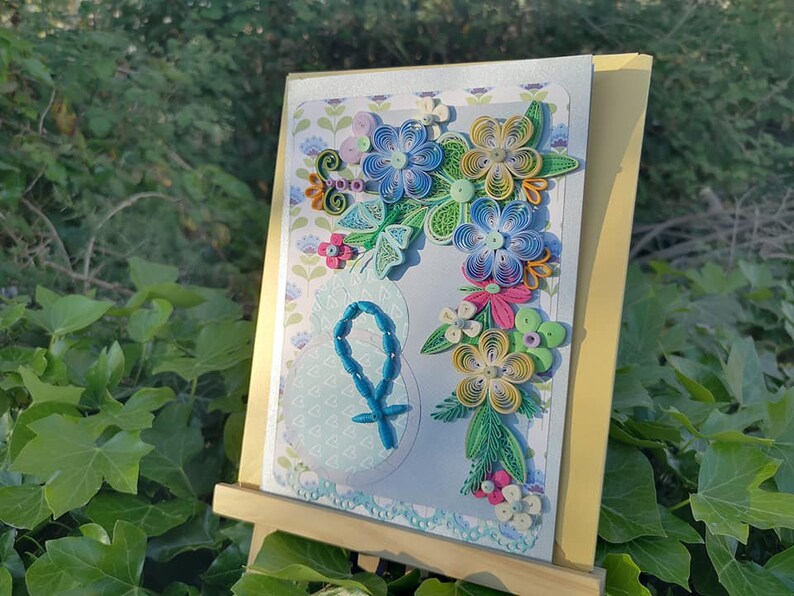 May include: A handmade card with a blue and green paper flower arrangement on a light blue background. The card features a blue rosary and a cross.