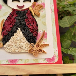 May include: A paper quilling art piece of a doll with a red, black, and white dress and a gold flower. The doll is on a green and white background with a red border.