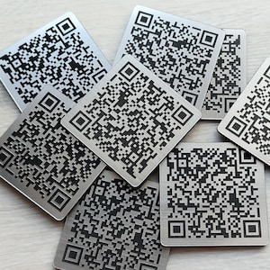 May include: A collection of square, metallic QR code tags. Each tag features a unique black and white matrix pattern within a square frame. The tags are arranged in a slightly overlapping pattern, showcasing their design and potential functionality.